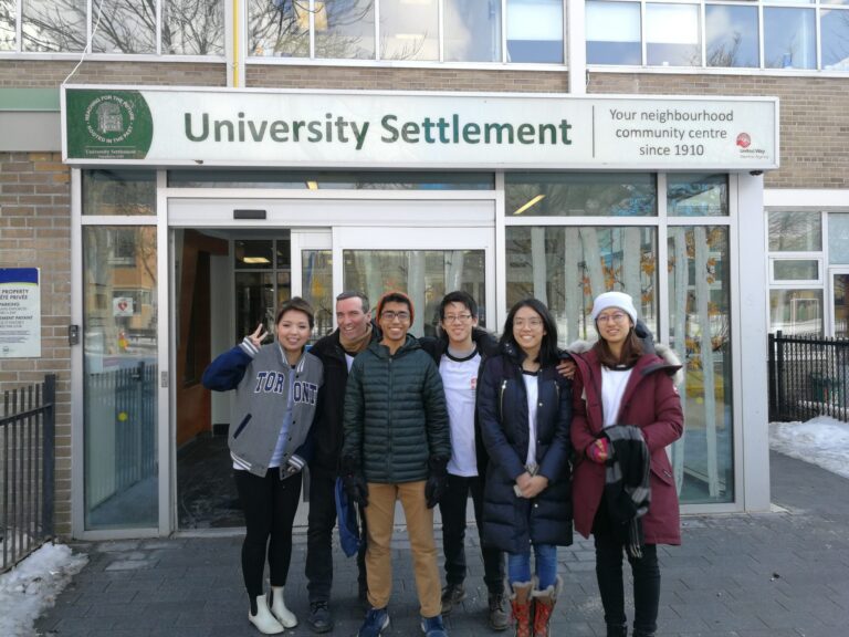 Six people stand in a line in front of a building with a sign that reads: University Settlement, Your neighbourhood community centre since 1910.