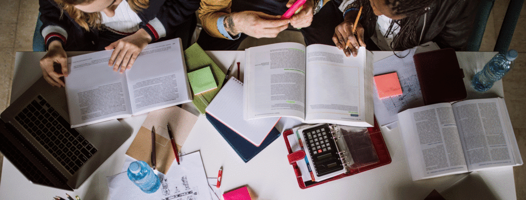 An overhead shot of three people seated at a table covered in books and devices.