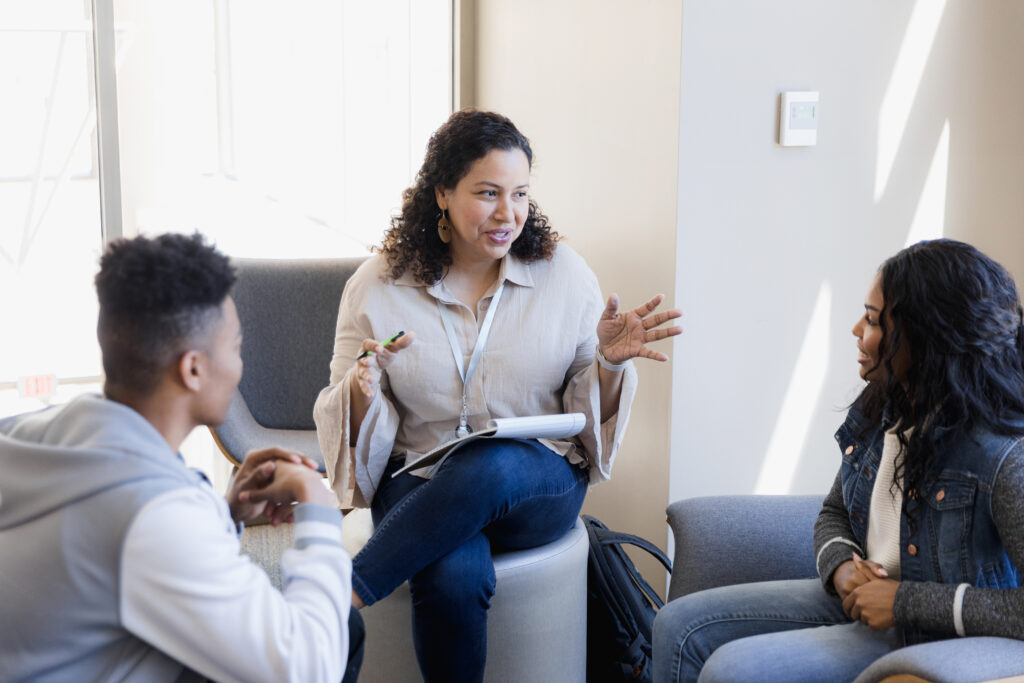 Two seated people listen intently to a person seated on a stool, speaking and holding a notepad on their lap.