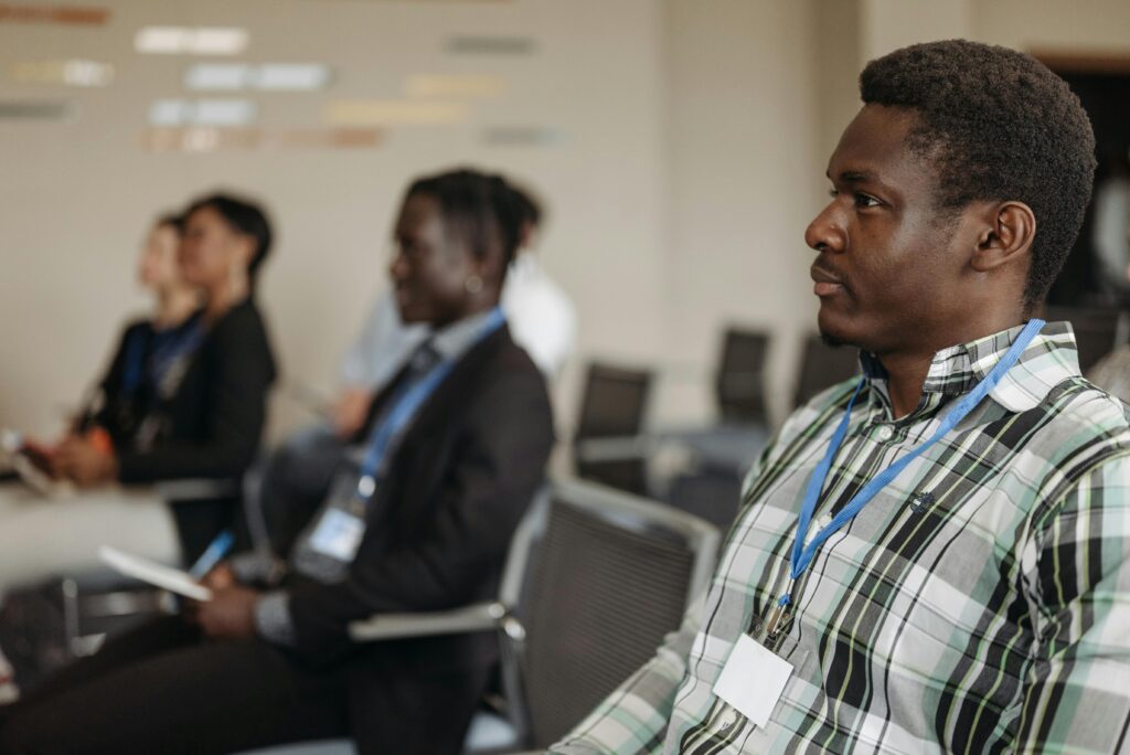 A Black man in focus, in a room of people who are seated, listens to a speaker (not pictured) at the front of the room.