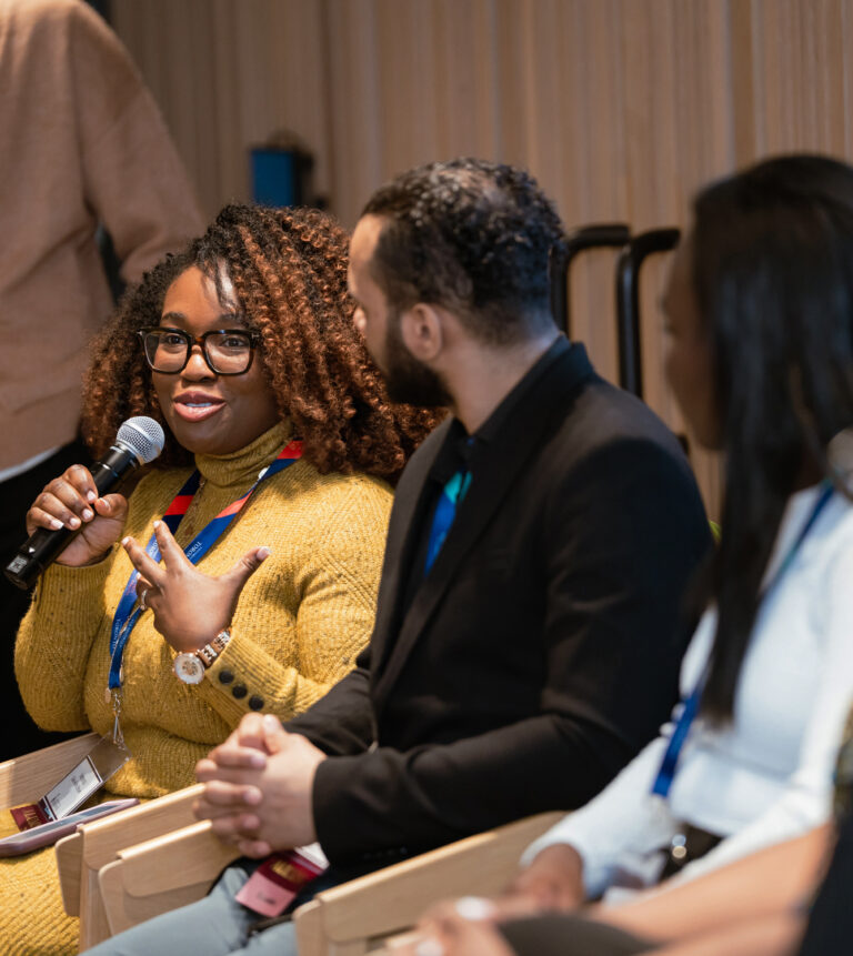 A Black person, on a panel with two other people in view, holds a microphone and speaks to the room.