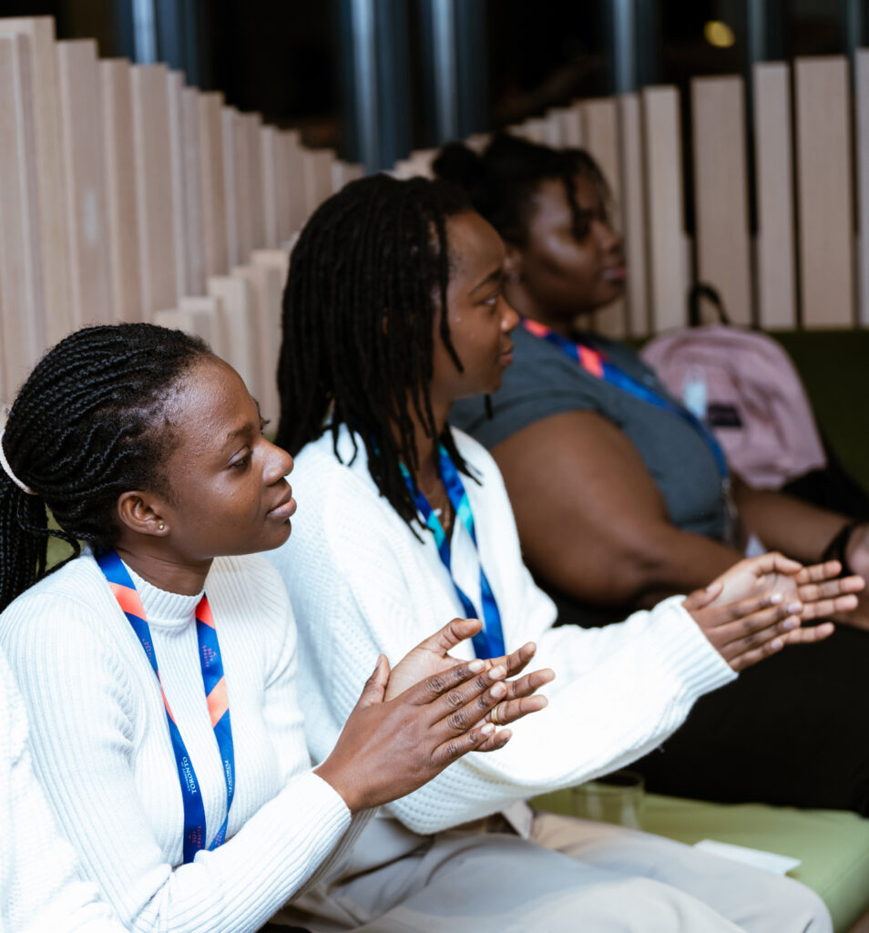 Three black people sit in a row, with lanyards on. They are looking towards the front of the room.