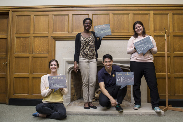 Four people pose in front of a fireplace and hold up signs. The signs read: I want to be an Entrepreneur, I want to be an Architect, I want to be a Professor and I want to make a difference.