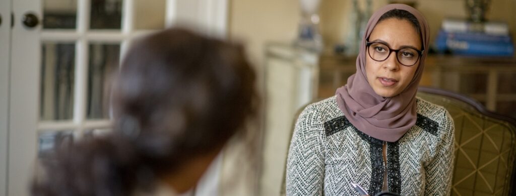 Two people site across from each other at a table. One person has their back to the camera and is out of focus. The other is a person, in focus, wearing a hijab and listening intently.