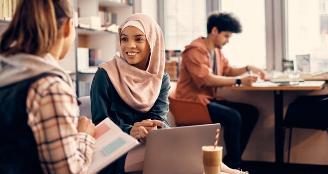 A person wearing a hijab and sitting with a laptop speaks to another person holding a notebook in a cafe. A third person sits at a table, writing, in the background.