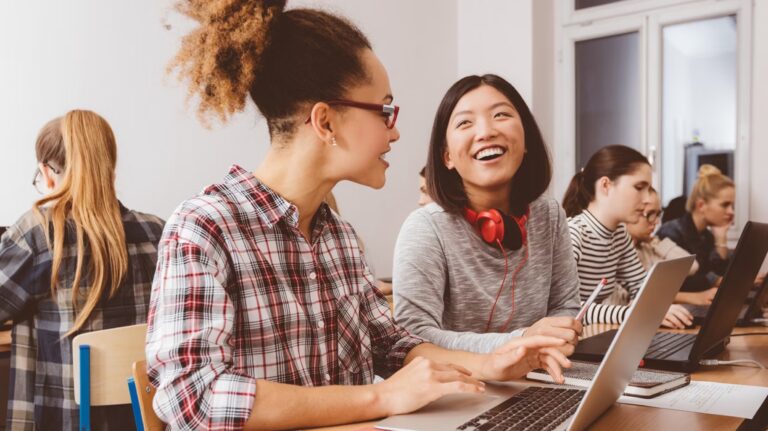 A group of people in a classroom, seated, with laptops. Two people are in focus, turned toward each other and smiling.