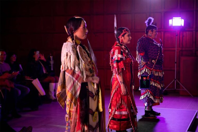 Three indigenous women model Indigenous Regalia on a runway.