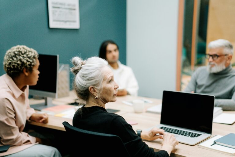 A group of four professionals seated around a conference room table with notebooks, laptops and computers. Focus on a woman looking intently to the front of the room