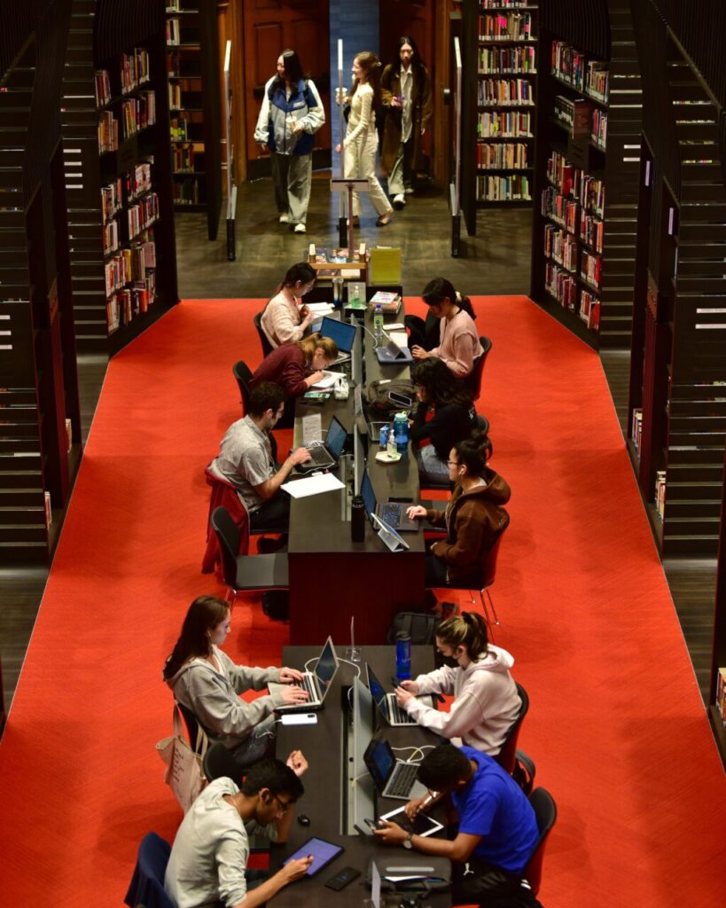 Many students seated and studying in a row of library study desks.