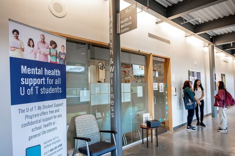 The front entrance to the Health and Wellness Centre framed by a banner that reads: Mental health supports for all U of T students and a group three students standing and chatting.