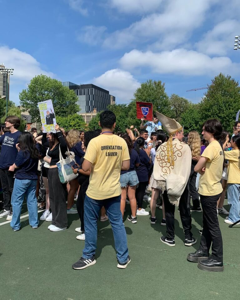 A group of students gathered for U of T's cheer off, carrying signs and wearing costumes. Focus on the back of one student who is wearing a yellow shirt that says Orientation Leader.