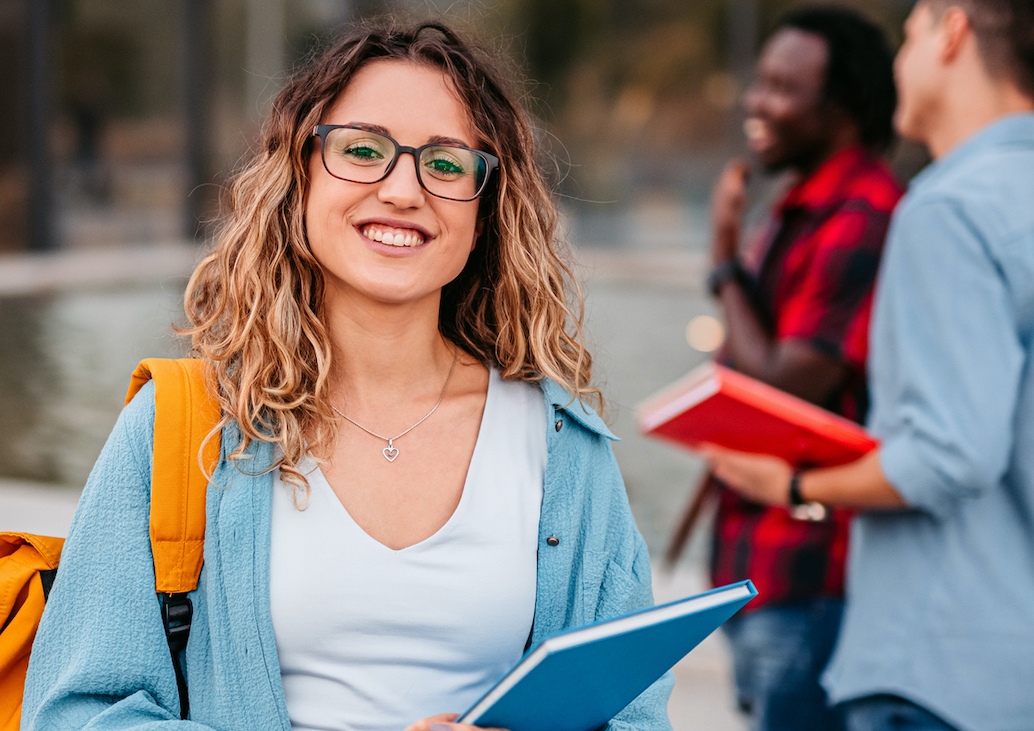 Portrait of a smiling university student standing on campus with friends in the background.