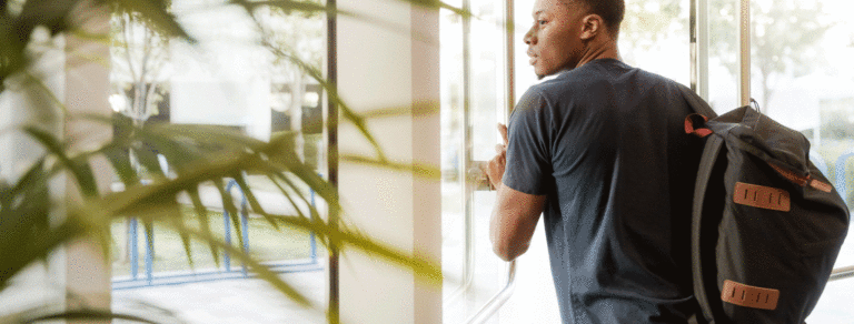 A young man leaves a building through a set of glass doors with a backpack on.