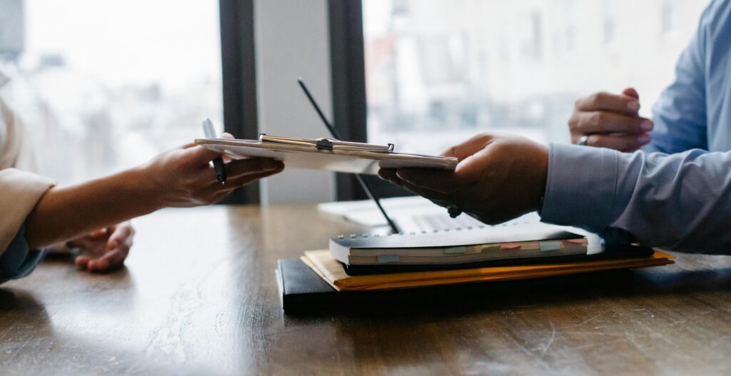 Two people pass a clipboard to each other across a table. Focus is on the hands of the people and the clipboard.