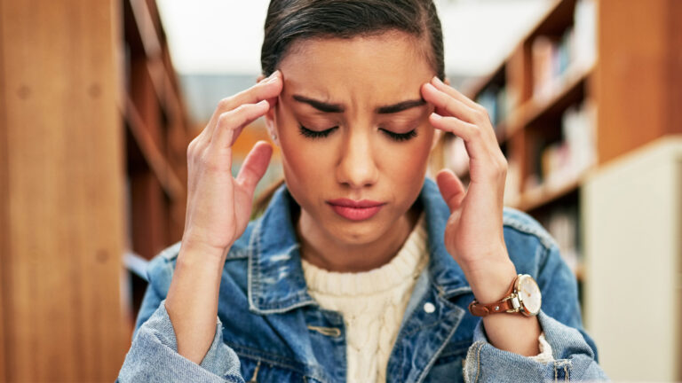 Close up of a person sitting at a table, looking stressed, massaging their temples with bookshelves in the background.