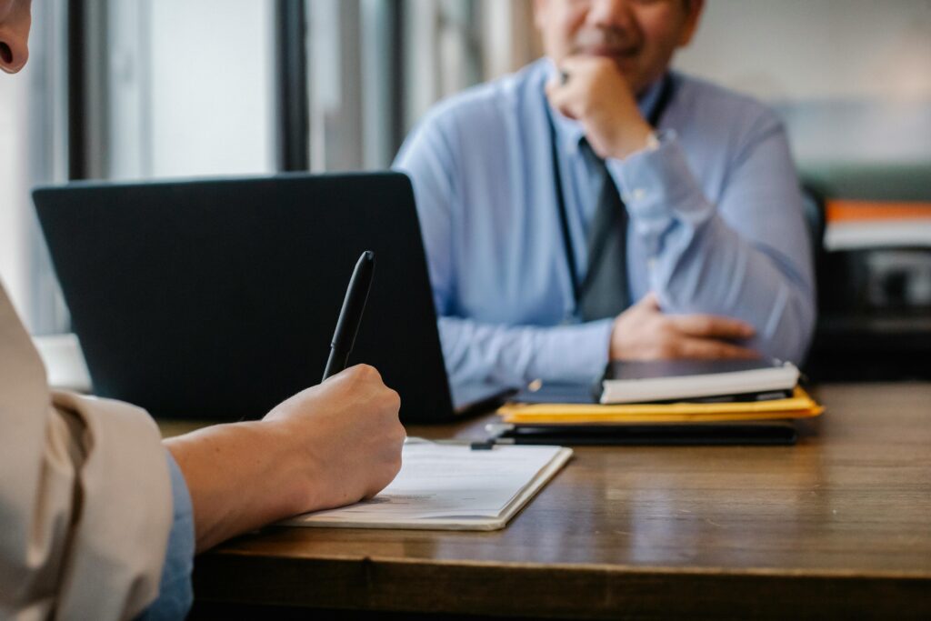 Two people sit across from each other at a desk with a laptop and pile of papers between them. Focus is on the hands of the person in the foreground, who is writing on a sheet of paper on a clipboard.