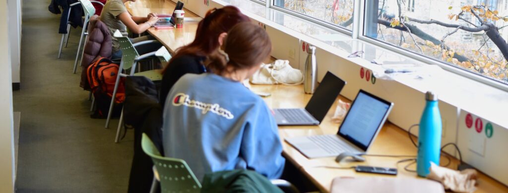 Two people sit at a library window table with their backs to the camera. They are looking at their laptop screens together.