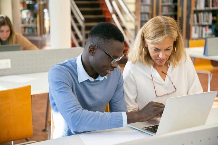 Two people sit together at a desk, looking at a computer screen in a library.