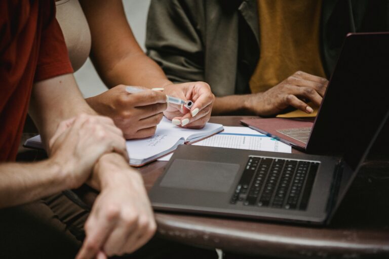 Three people sit around a table in front of a computer. One person has a notebook and pen in hand