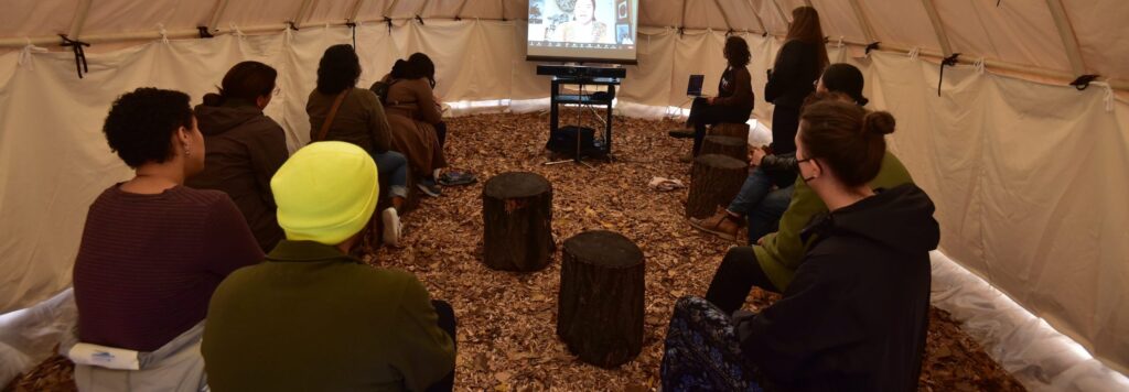 Group of people sitting on logs in a tipi watching a screen