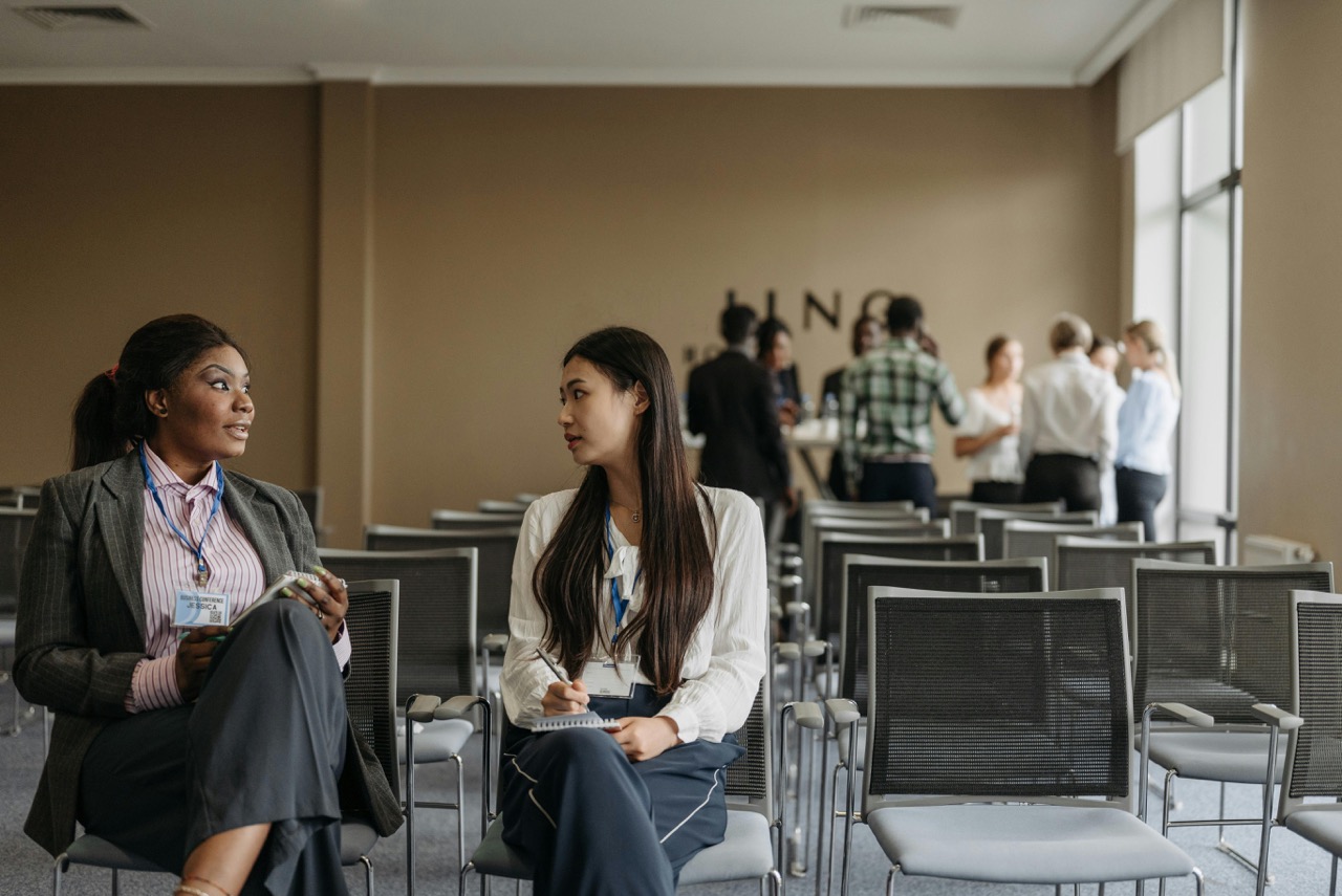 Two women speaking to each other sitting in chairs at the front of a conference room